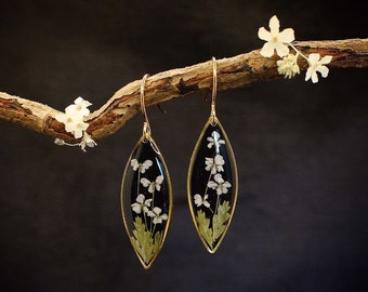 May include: Gold earrings with delicate white flowers pressed inside clear resin, hanging from a branch with white flowers.
