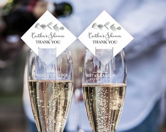 May include: Two champagne flutes with a white diamond-shaped thank you card with green foliage and the text "Caitlin's Shower Thank You" on each.
