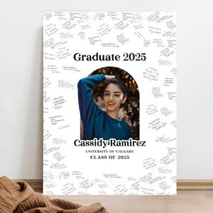 May include: A white graduation sign with black text that reads "Graduate 2025" and "Cassidy Ramirez, University of Calgary, Class of 2025". A photo of a woman with dark hair and a blue sweater is in a circular cutout in the sign. The sign is covered in black ink signatures.