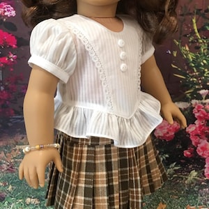 May include: A white blouse with a lace trim and buttons, paired with a brown and tan plaid skirt. The doll is wearing a beaded bracelet.