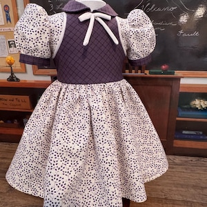 May include: A vintage-style dress with a purple bodice and a white bow. The dress features a white and purple floral pattern on the skirt and sleeves. The bodice has a diamond pattern. The dress is displayed on a mannequin.