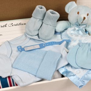 May include: A blue baby gift set in a white box. The set includes a blue onesie, a blue hat, a pair of blue booties, a blue teddy bear, and a blue blanket. The box also includes a birth certificate card.