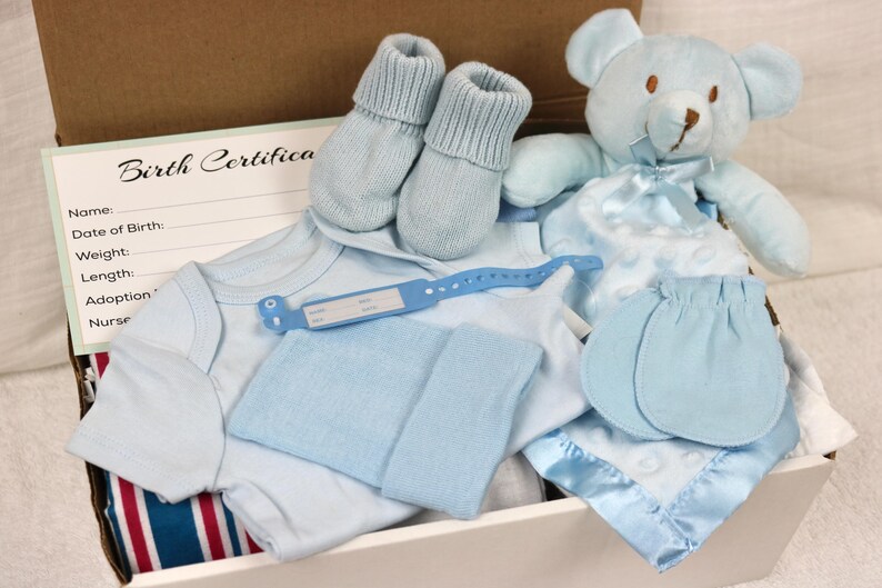 May include: A blue baby gift set in a white box. The set includes a blue onesie, a blue hat, a pair of blue booties, a blue teddy bear, and a blue blanket. The box also includes a birth certificate card.