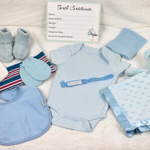 May include: A blue baby outfit set with a onesie, hat, booties, mittens, bib, and a blue blanket with a teddy bear. The set is laid out on a white surface with a birth certificate card.