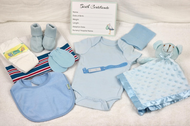 May include: A blue baby outfit set with a onesie, hat, booties, mittens, bib, and a blue blanket with a teddy bear. The set is laid out on a white surface with a birth certificate card.
