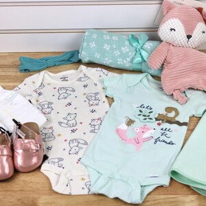 May include: A baby gift set with a green and white patterned blanket, a white onesie with a fox print, a light blue onesie with a fox and squirrel print, a pink fox plush toy, a pair of pink baby shoes, and a white headband with a green bow.