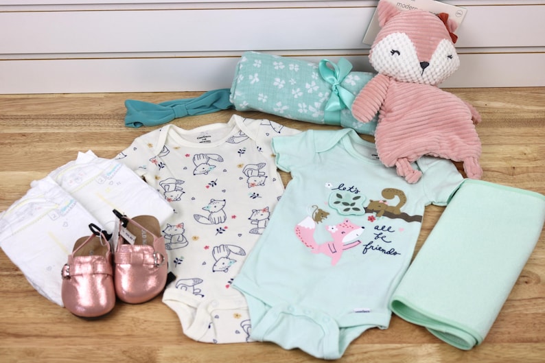 May include: A baby gift set with a green and white patterned blanket, a white onesie with a fox print, a light blue onesie with a fox and squirrel print, a pink fox plush toy, a pair of pink baby shoes, and a white headband with a green bow.