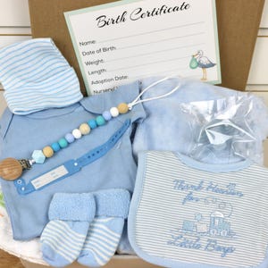 May include: A blue baby gift basket with a blue and white striped hat, a blue onesie, blue socks, a blue blanket, a blue bib with the text "Thank heavens for little boys", a wooden teether, and a blue and white beaded necklace.