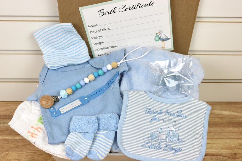 May include: A blue baby gift basket with a blue and white striped hat, a blue onesie, blue socks, a blue blanket, a blue bib with the text "Thank heavens for little boys", a wooden teether, and a blue and white beaded necklace.