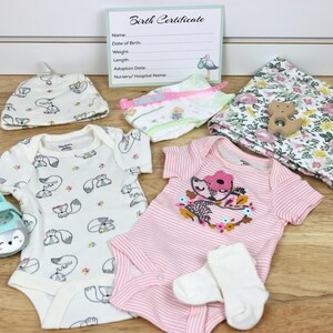 May include: A baby gift set with a pink and white striped onesie, a white onesie with a fox print, a white hat with a fox print, a floral print blanket, a pair of white socks, a pair of blue shoes, and a wooden toy.