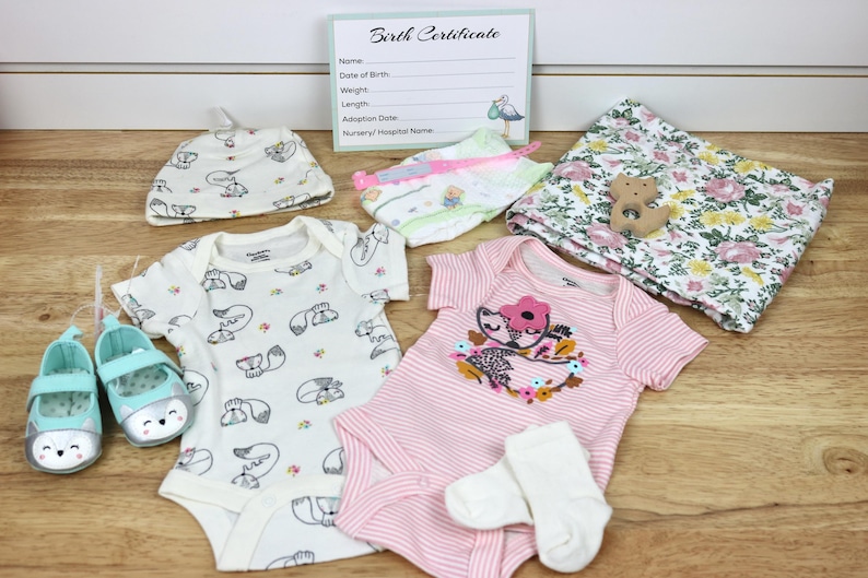 May include: A baby gift set with a pink and white striped onesie, a white onesie with a fox print, a white hat with a fox print, a floral print blanket, a pair of white socks, a pair of blue shoes, and a wooden toy.