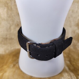 May include: A black suede bracelet with a silver-colored buckle is displayed on a white mannequin. The bracelet has a simple, classic design, with a single strap and a buckle closure. The background is a soft, golden color.
