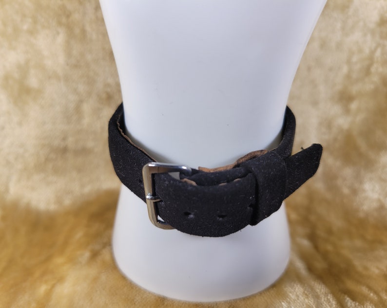 May include: A black suede bracelet with a silver-colored buckle is displayed on a white mannequin. The bracelet has a simple, classic design, with a single strap and a buckle closure. The background is a soft, golden color.