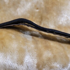 May include: A close-up view of a black, curved, elastic band, possibly for a headband or hair accessory. The band is layered, with a lighter-colored material visible beneath the black exterior. The background is a soft, textured, golden-yellow fabric.