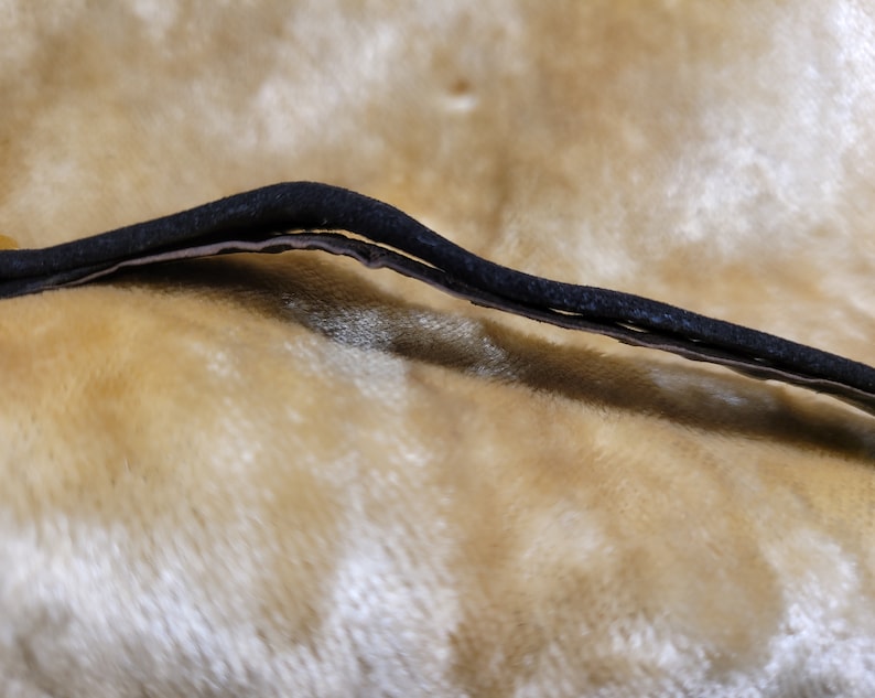 May include: A close-up view of a black, curved, elastic band, possibly for a headband or hair accessory. The band is layered, with a lighter-colored material visible beneath the black exterior. The background is a soft, textured, golden-yellow fabric.