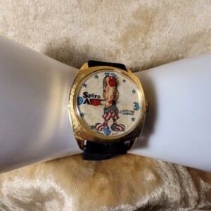 May include: A vintage gold-toned watch with a black band. The watch face features a cartoon character in red, white, and blue, with the words "Spiro Agnew" and "Time Unlimited" printed on the face. The watch is on a white surface.