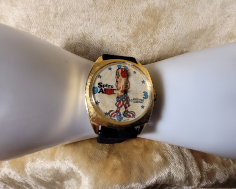 May include: A vintage gold-toned watch with a black band. The watch face features a cartoon character in red, white, and blue, with the words "Spiro Agnew" and "Time Unlimited" printed on the face. The watch is on a white surface.
