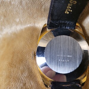 May include: A gold-toned wristwatch with a black leather band. The watch has a round face with a silver-colored back. The back of the watch has the text "STAINLESS STEEL SWISS" and the number "13323".