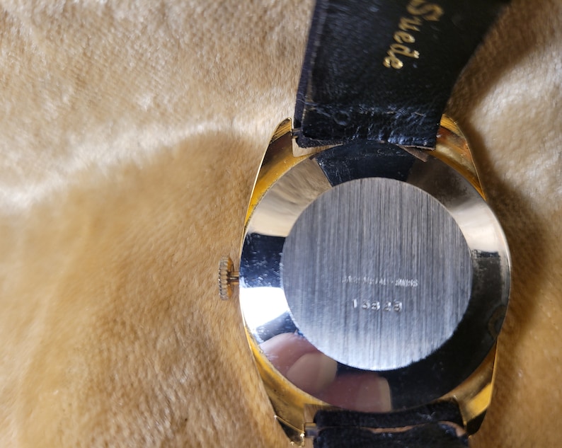 May include: A gold-toned wristwatch with a black leather band. The watch has a round face with a silver-colored back. The back of the watch has the text "STAINLESS STEEL SWISS" and the number "13323".