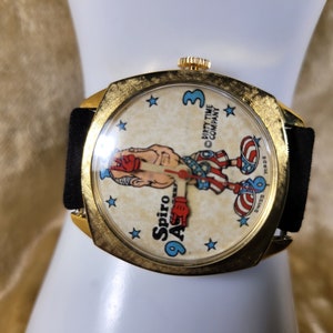 May include: A vintage gold-toned wristwatch with a black band. The watch face features a cartoon boxer with red gloves and patriotic attire. The text "Spiro Agnew" and "Dirty Time Company" are visible.
