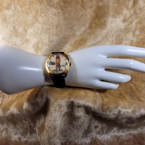 May include: Vintage gold-toned watch with a black leather band. The watch face features a cartoon character and the text "Spiro ABC". The watch is displayed on a white mannequin hand.