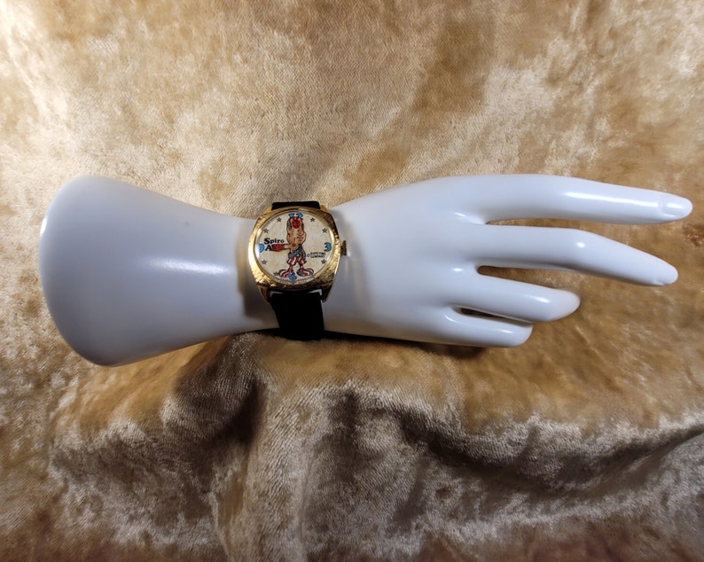 May include: Vintage gold-toned watch with a black leather band. The watch face features a cartoon character and the text "Spiro ABC". The watch is displayed on a white mannequin hand.