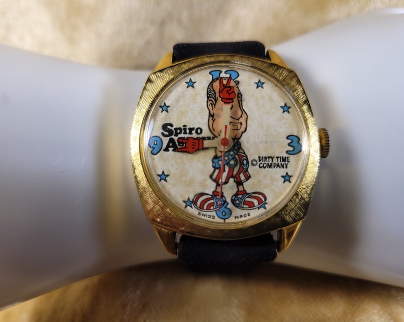 May include: Vintage gold-tone watch with a black band. The watch face features a cartoon character with the text "Spiro Agnew" and "Dirty Time Company". The watch has a textured bezel and a Swiss Made label.