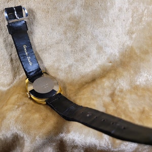May include: A gold-toned wristwatch with a black leather band. The watch face is not visible. The band is labeled "Genuine Suede". The watch is set against a light brown, textured background.