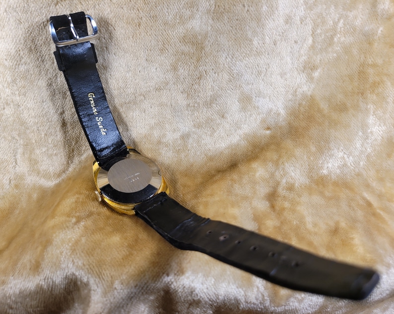 May include: A gold-toned wristwatch with a black leather band. The watch face is not visible. The band is labeled "Genuine Suede". The watch is set against a light brown, textured background.
