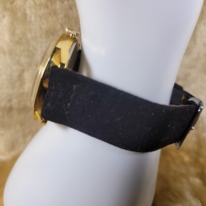 May include: A gold-toned wristwatch with a black leather band is displayed. The watch face is round with a gold bezel. The black band is secured with a silver buckle. The watch is presented on a white mannequin hand.