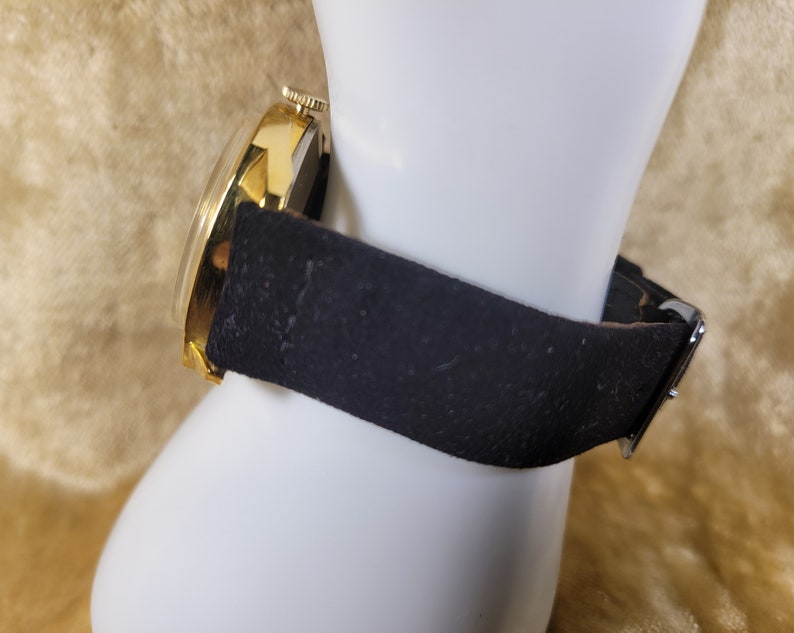 May include: A gold-toned wristwatch with a black leather band is displayed. The watch face is round with a gold bezel. The black band is secured with a silver buckle. The watch is presented on a white mannequin hand.