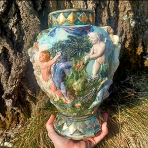 Op de afbeelding: Een gedetailleerde, handgeschilderde keramische vaas met een turquoise, gouden en groene kleurstelling. De vaas heeft reliëfsculpturen van figuren in een tuin. De basis en rand hebben een ruitpatroon.