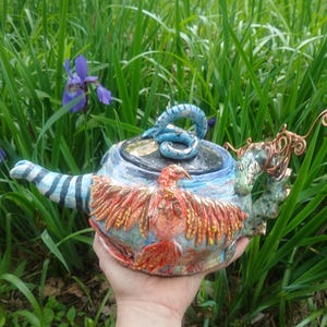 Op de afbeelding: Een handgemaakte keramische theepot met een feniksontwerp. De theepot heeft een blauw-wit gestreepte handgreep, een blauwe slangvormige dekselgreep en een rode, oranje en gele feniks op de body. De tuit heeft een groen draakachtig ontwerp.