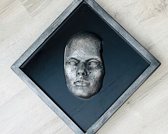 May include: A silver-colored face sculpture is encased in a black resin frame with a silver-colored border. The sculpture is set against a black background.