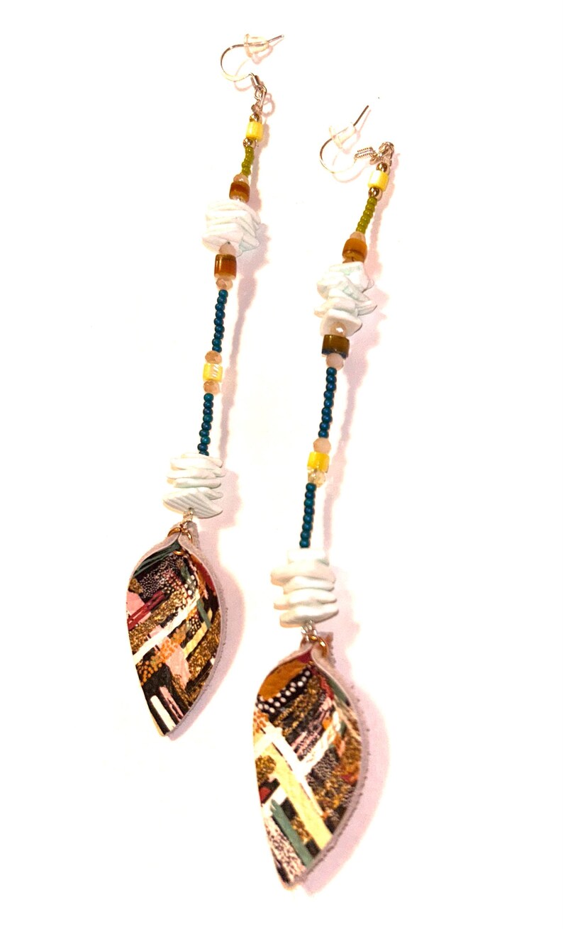 Puede incluir: Pendientes colgantes largos con un colgante en forma de hoja. Los pendientes presentan una combinaci&oacute;n de cuentas blancas, marrones, amarillas y verdes. Los colgantes de hoja tienen un dise&ntilde;o de patchwork colorido con detalles dorados.
