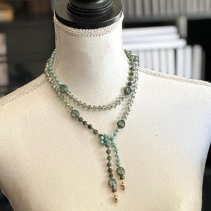 May include: A layered necklace with a delicate design. The necklace features a combination of small, clear beads and larger, round, teal-colored beads. The necklace is displayed on a white mannequin, showcasing its length and style. The necklace has a tassel-like drop.