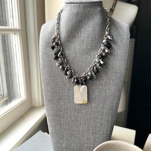 May include: A silver-toned necklace with a rectangular pendant and dark gray beads. The necklace is displayed on a gray mannequin. The pendant is a light cream color with a rectangular shape. The necklace is a statement piece.