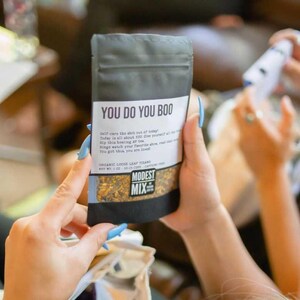 May include: A black and gray tea bag with the text "YOU DO YOU BOO" in white. The bag also includes the text "MODEST MIX" and other text. The bag is held by a person with blue fingernails.