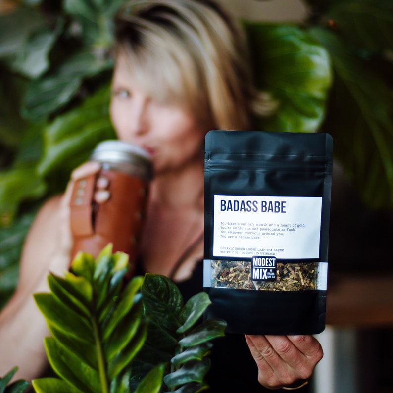 May include: A black package of "BADASS BABE" organic green loose leaf tea blend. The package has white text and a small logo. The image also shows a woman holding the tea package and a mason jar with a brown leather sleeve.