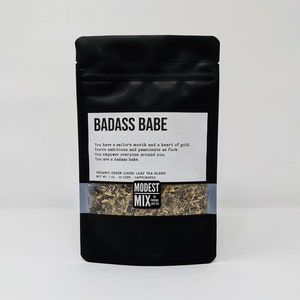 May include: Black pouch of "BADASS BABE" organic green loose leaf tea blend. The label includes text about being ambitious and passionate. The bottom of the pouch shows the tea leaves. Net weight is 2 oz, about 20 cups.