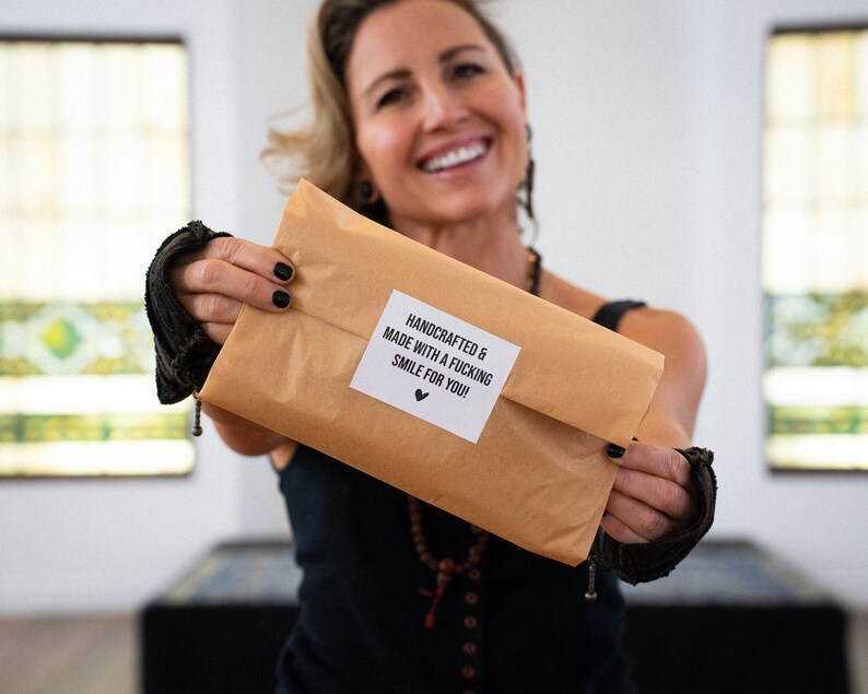 May include: A brown paper package with a white sticker that reads "HANDCRAFTED & MADE WITH A FUCKING SMILE FOR YOU!" with a heart symbol. The package is being held up by a person.