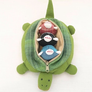 May include: A green turtle-shaped soft toy with a plaid pattern. The turtle's shell opens with a zip to reveal three small, round, smiling dolls in red, black, and blue. The turtle has a tail, four flippers, and a smiling face.