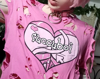 Könnte beinhalten: Pinkes T-Shirt mit einem großen Herz-Motiv und dem Text "Succuboi" in rosa-weißer Schrift. Das Shirt ist zerrissen und abgenutzt mit Löchern überall. Die Person, die das Shirt trägt, trägt auch rosa Katzenohren und eine rosa Halskette.