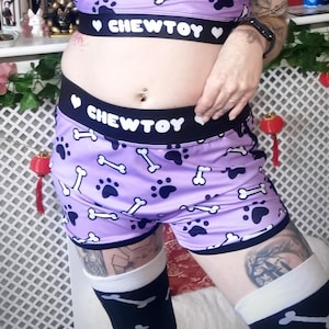 May include: A lavender two-piece set featuring a paw print and bone design. The set includes a crop top and shorts, both with the word "CHEWTOY" printed on the waistband. The person is wearing black and white thigh-high socks with a bone design.