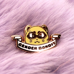 May include: A gold-coloured enamel pin featuring a cartoon cat wearing a black mask and a white banner that reads "Gender Bandit".