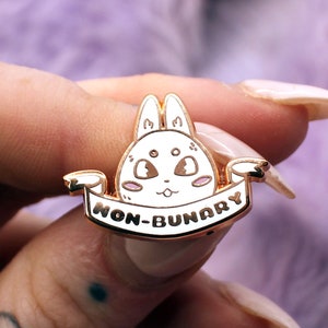 Peut inclure: Épinglette en émail doré représentant un lapin blanc de dessin animé avec des joues roses et le texte "NON-BINAIRE" sur une bannière.