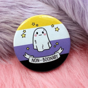 May include: A round button featuring a non-binary pride flag design. The flag is yellow, white, purple, and black. A cartoon ghost is in the centre of the flag. The text "NON-BINARY" is on a banner below the ghost.