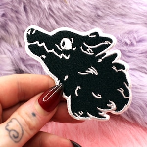 May include: A black and white embroidered wolf head patch with a white border. The patch is set against a backdrop of soft, fluffy, pink and lilac material. The design is detailed.