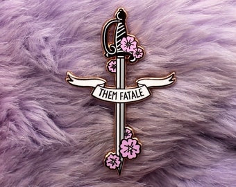 Può includere: Uno spillo in smalto dorato con una spada con fiori rosa e uno stendardo che recita "Them Fatale".