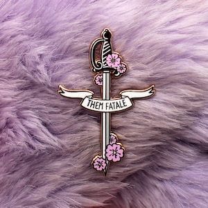 May include: A gold-toned enamel pin featuring a sword with pink flowers and a banner that reads "Them Fatale".